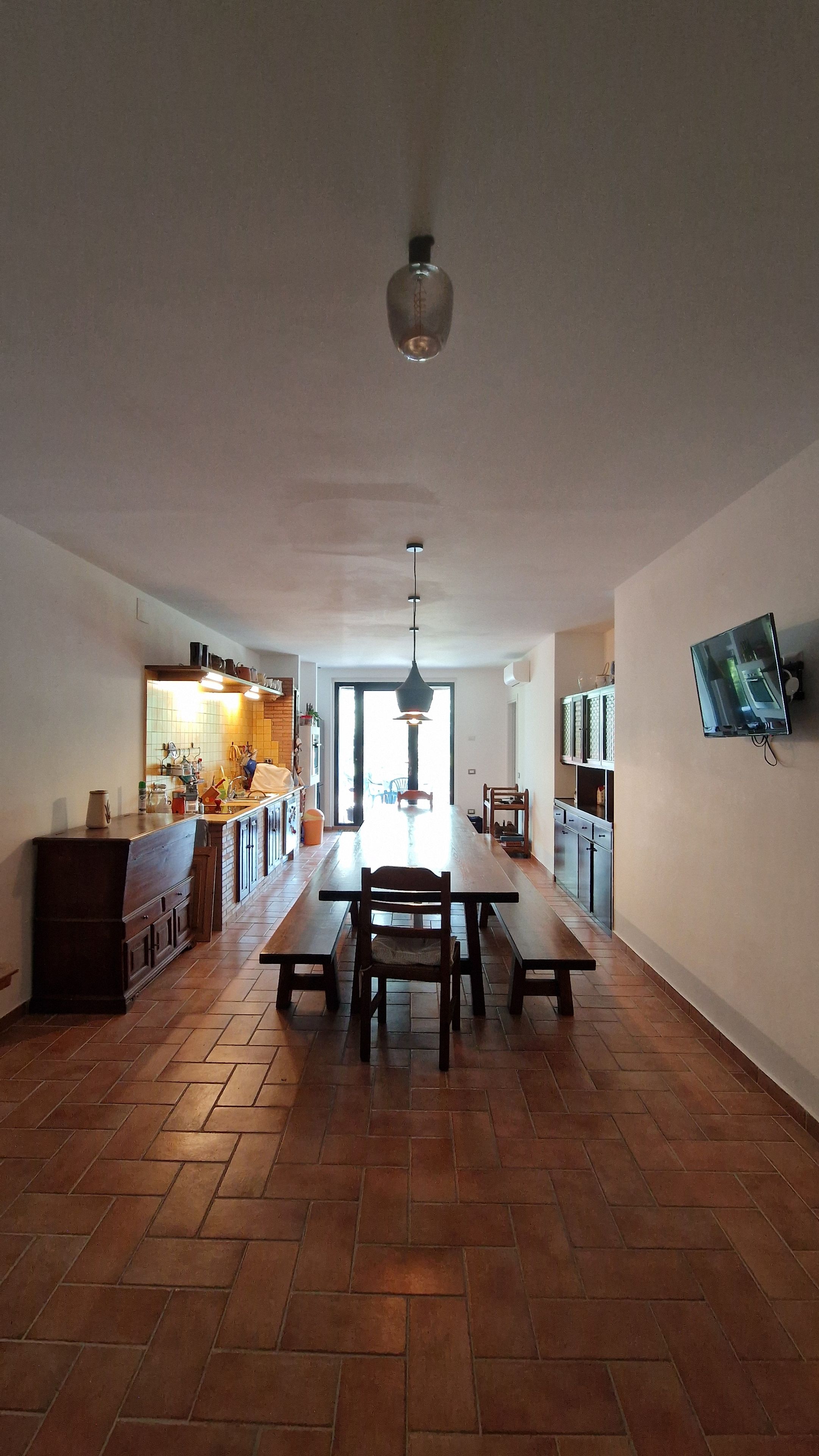 A spacious kitchen with a dining area, a TV mounted on the wall, and a large window.