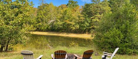 Three Adirondack chairs facing a lake surrounded by trees.
