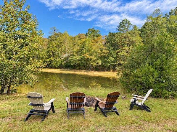 Three Adirondack chairs facing a lake surrounded by trees.