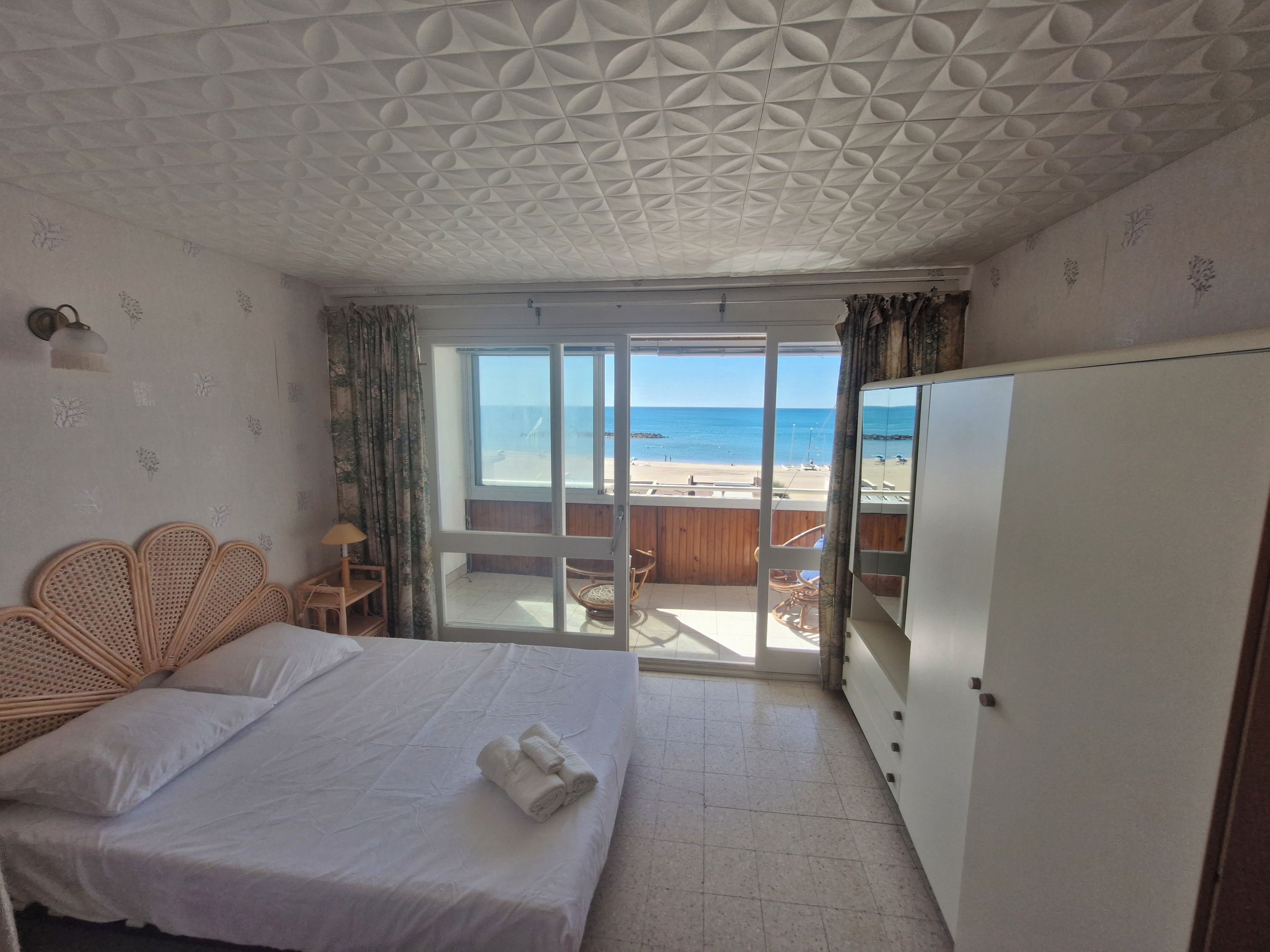 A hotel room with a bed, a wardrobe, a balcony with a view of the beach, and a patterned ceiling.