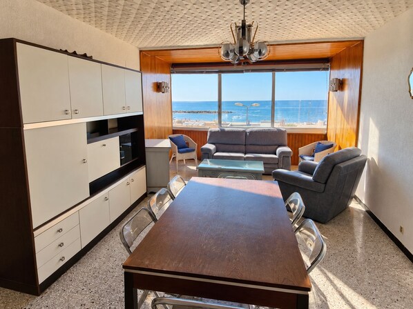 Un espace de vie compact comprenant un coin cuisine, une table à manger et un coin salon. Une grande fenêtre offre une vue sur la plage.