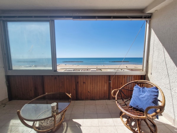 A balcony with a view of the beach, a glass table, and a wicker chair. - Vrbo Property (Valras-Plage)