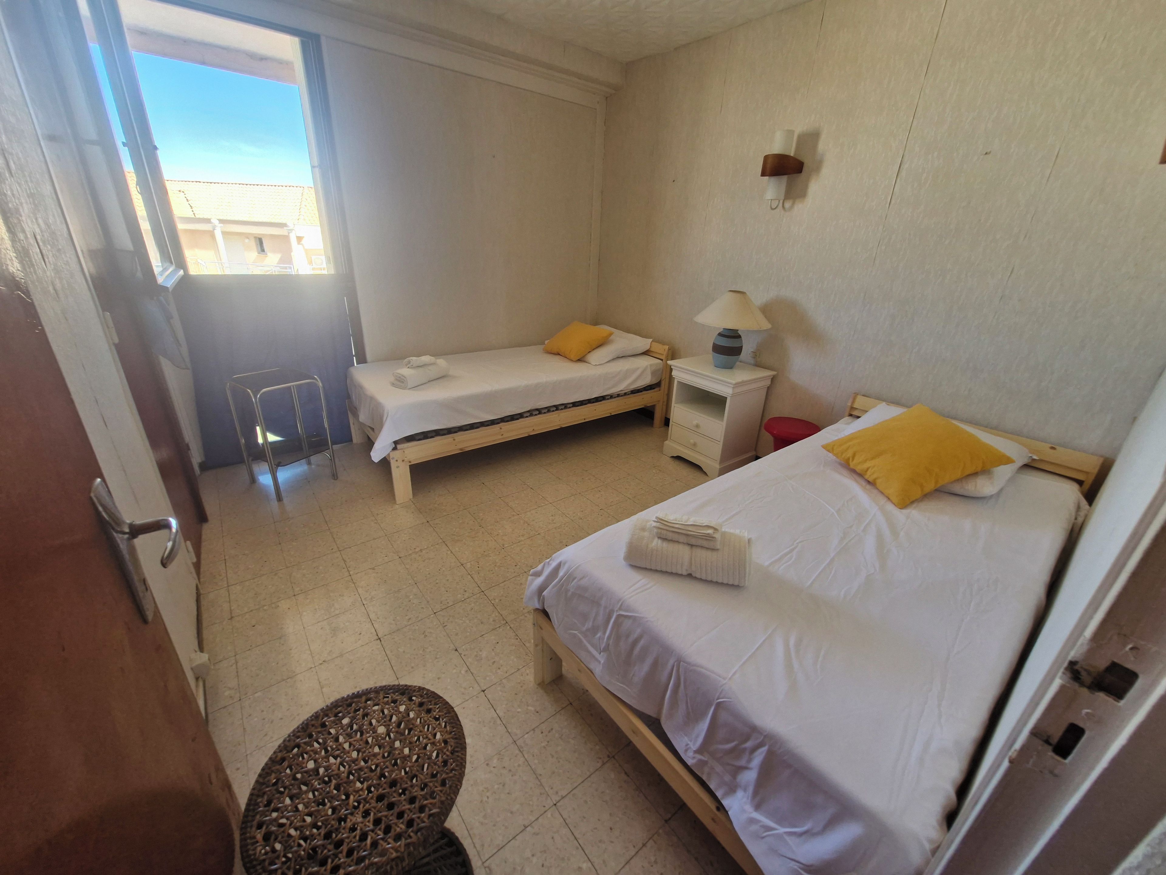 A hotel room with two single beds, a small table with a lamp, a chair, and a window with a view of buildings outside.