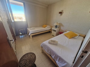 A hotel room with two single beds, a small table with a lamp, a chair, and a window with a view of buildings outside. - Vrbo Property (Valras-Plage)