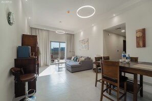 A modern living room with a sofa, dining table, and chairs. There is a large window with curtains, a painting on the wall, and a view of the cityscape. - 2BR + Huge Private Terracen- Al Furjan Near Metro (Dubai)
