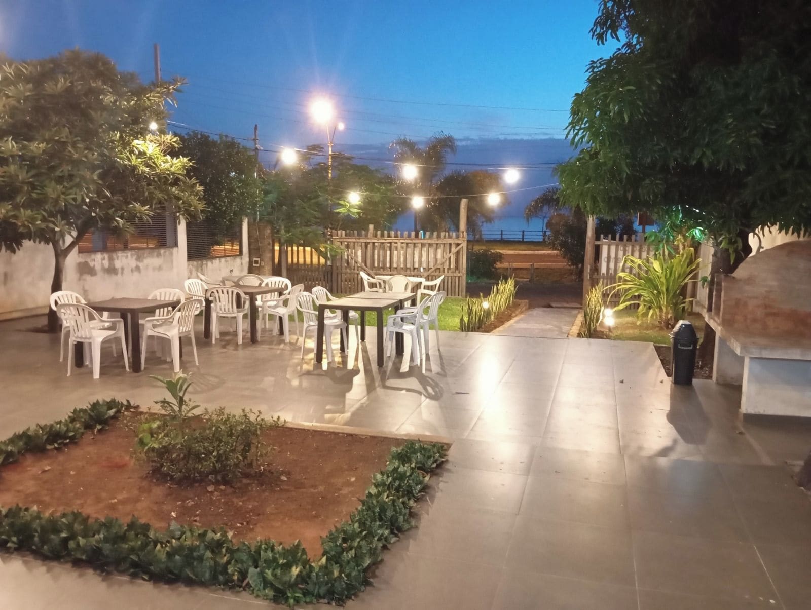 An outdoor patio area with tables and chairs, surrounded by plants and a wooden fence.