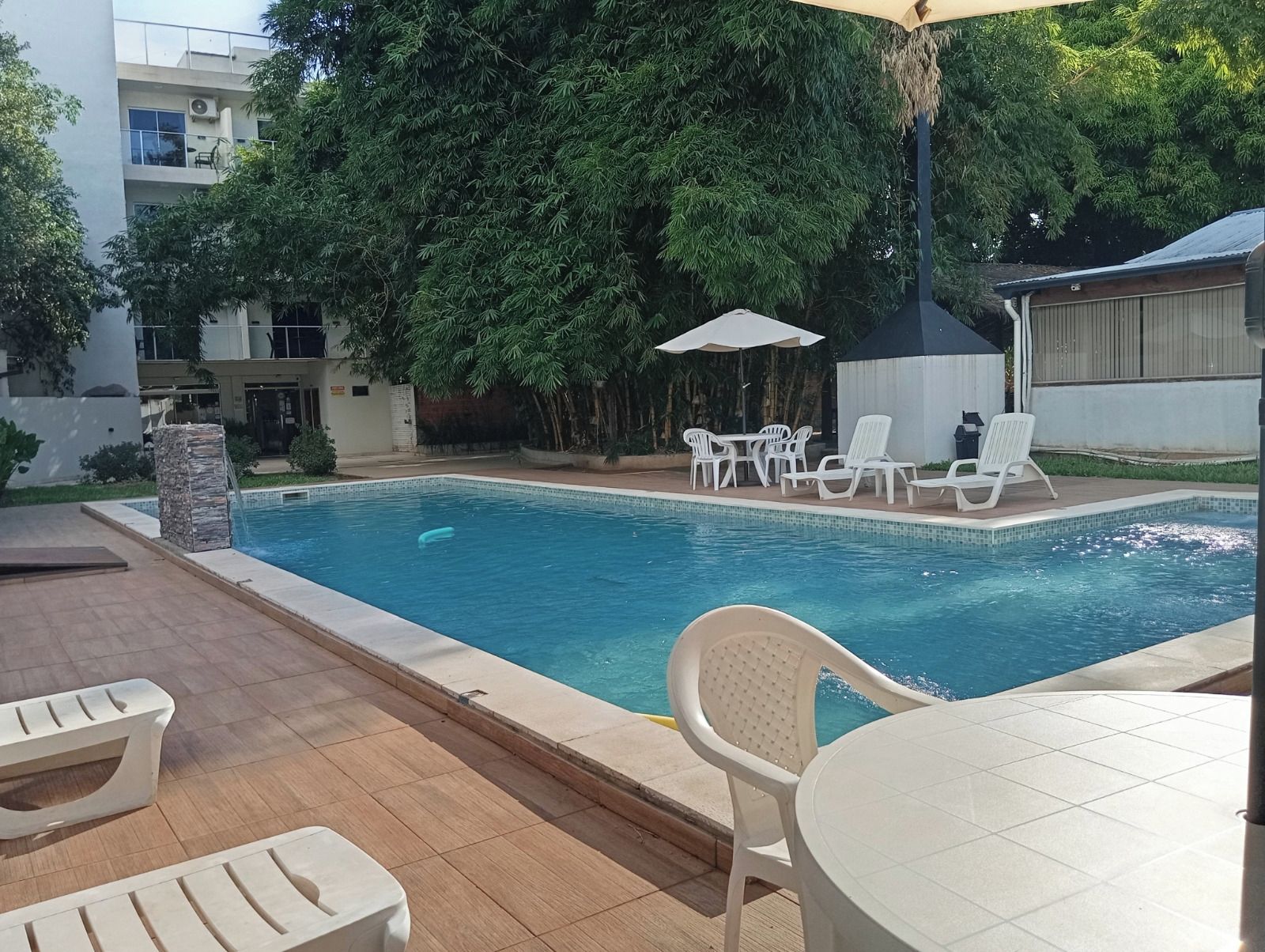 Una piscina con sillones y mesas, rodeada de vegetación y un edificio al fondo.