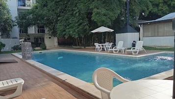 Una piscina con sillones y mesas, rodeada de vegetación y un edificio al fondo.