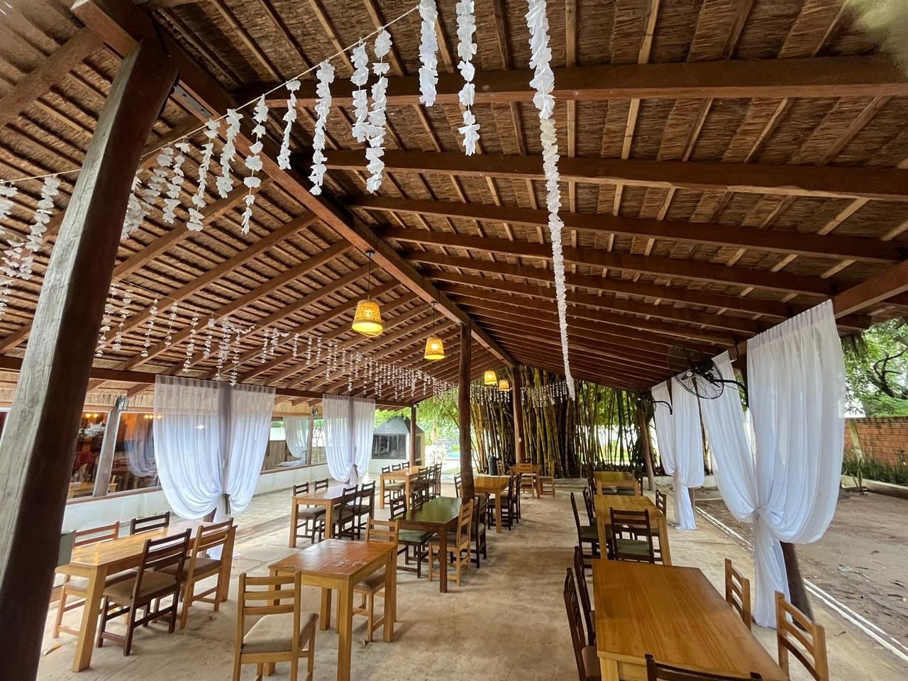 An outdoor dining area with wooden tables and chairs, covered by a wooden roof with hanging decorations.