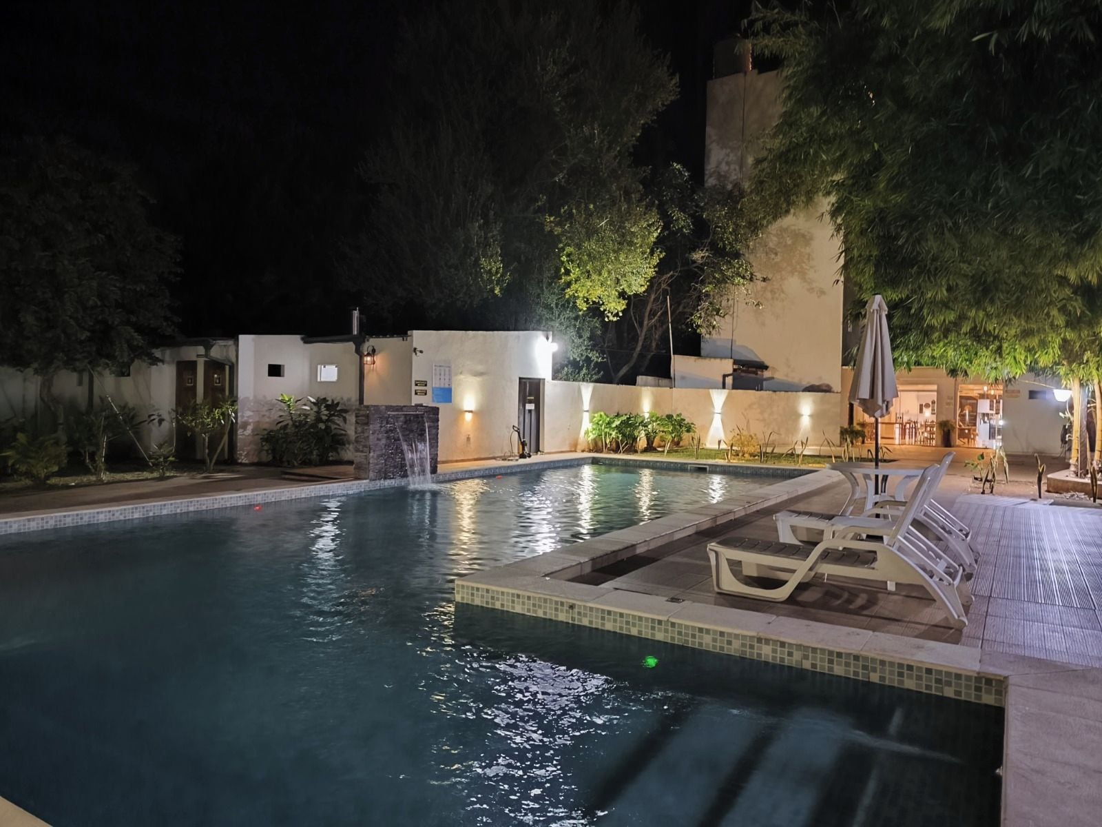 An outdoor swimming pool with a tiled edge, surrounded by a low wall and trees. There are lounge chairs and umbrellas on the poolside.