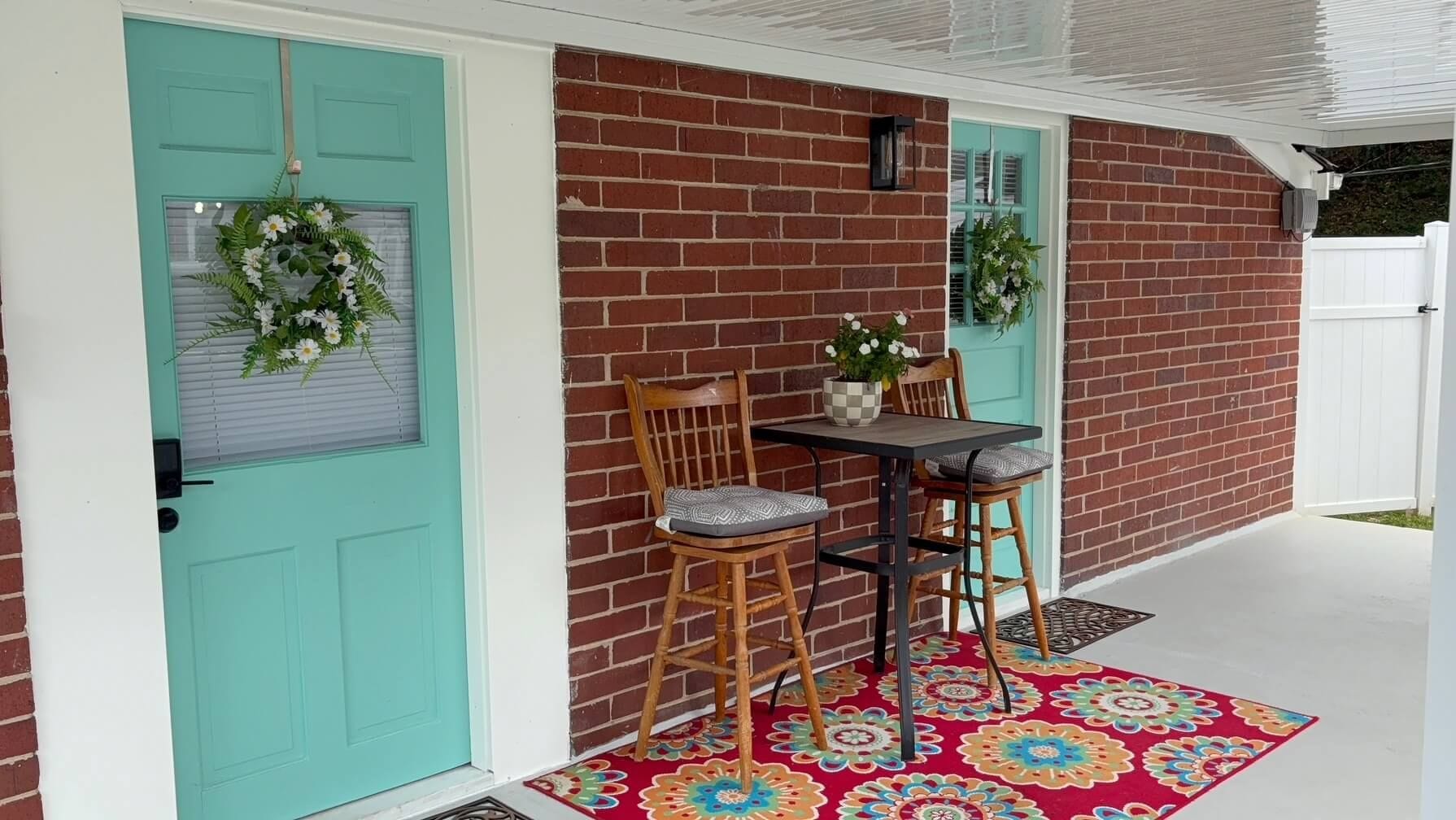 A porch with a teal door, a brick wall, a small table with two chairs, and a colorful rug.
