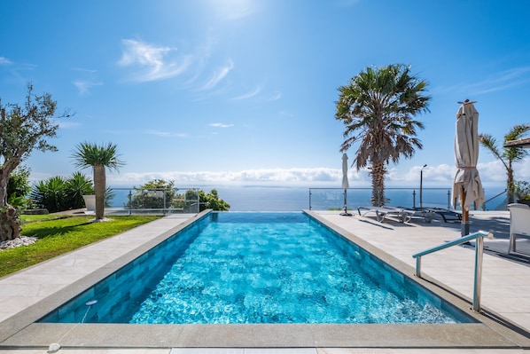 A rectangular swimming pool with clear blue water, surrounded by a tiled deck. There are palm trees and a view of the ocean in the background. - Vrbo Property (Fajã da Ovelha)