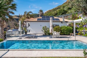 A swimming pool with a white building in the background, solar panels on the roof, and a hillside view. - Vrbo Property (Fajã da Ovelha)