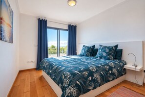 Een slaapkamer met een groot bed, een nachtkastje, een lamp en uitzicht op het landschap door een raam.