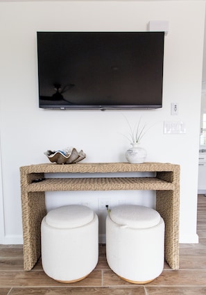 A flat-screen TV mounted on a white wall above a woven console table with two round ottomans beneath. - Walk to Roger Wheeler Beach - Water Views (Narragansett)