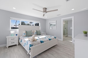 A bedroom with a bed, nightstands, a ceiling fan, a window with blinds, and a door leading to a bathroom. - Beachside Oasis (Fort Myers Beach)