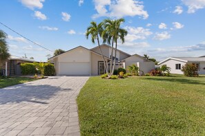 A single-story house with a garage, a paved driveway, and a well-maintained lawn with various plants and a palm tree. - Canal Paradise Retreat (Fort Myers Beach)