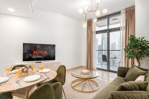 A modern living room with a dining area, a flat-screen TV, a large window with a city view, and a potted plant. - FIRST CLASS | 2BR | Burj View | Premium City Stay (Dubai)