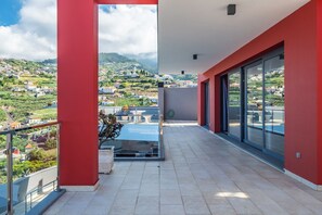 A balcony with a view of a hillside village, a potted plant, and a glass door leading to the interior. - Villa Rodrigues (Ponta do Sol)