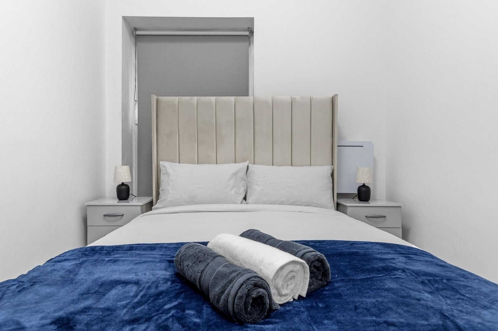 A bed with a headboard, two bedside tables with lamps, and a blue blanket with rolled towels.