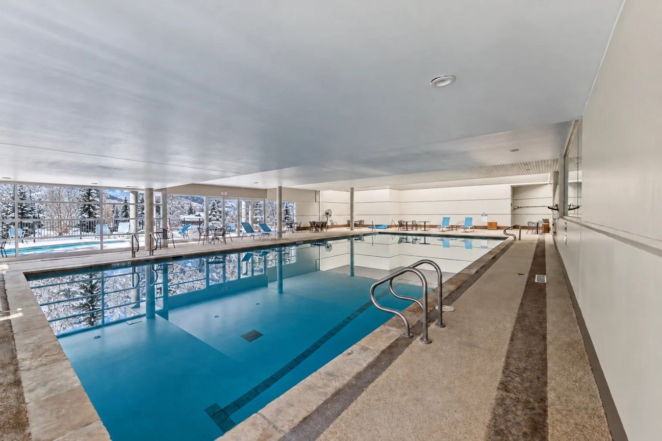 A large indoor swimming pool with clear glass walls, a view of snowy trees, and a modern interior design.