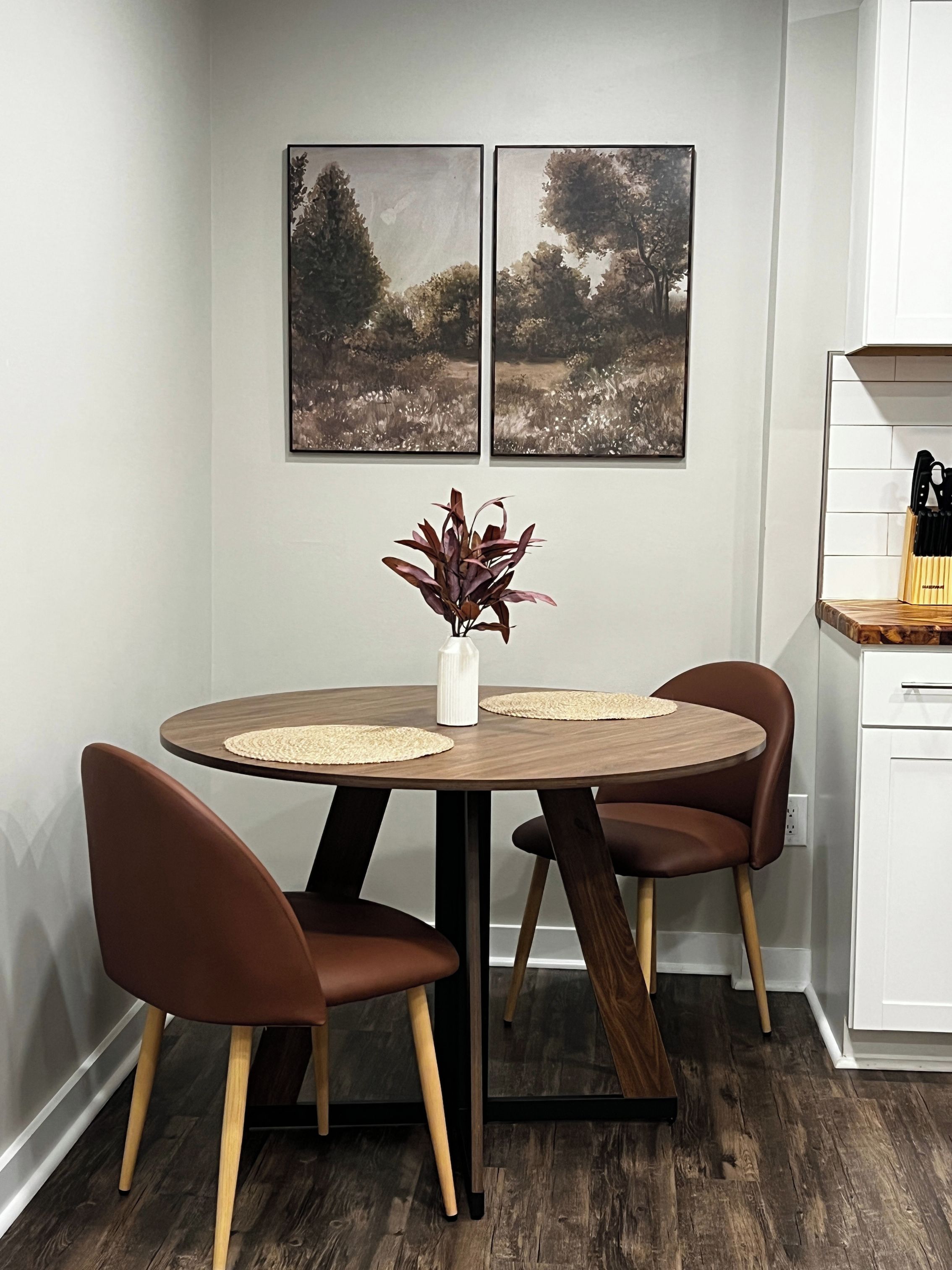Ein Essbereich mit einem runden Tisch, zwei Stühlen, einer Vase mit Pflanze und zwei gerahmten Bildern an der Wand.