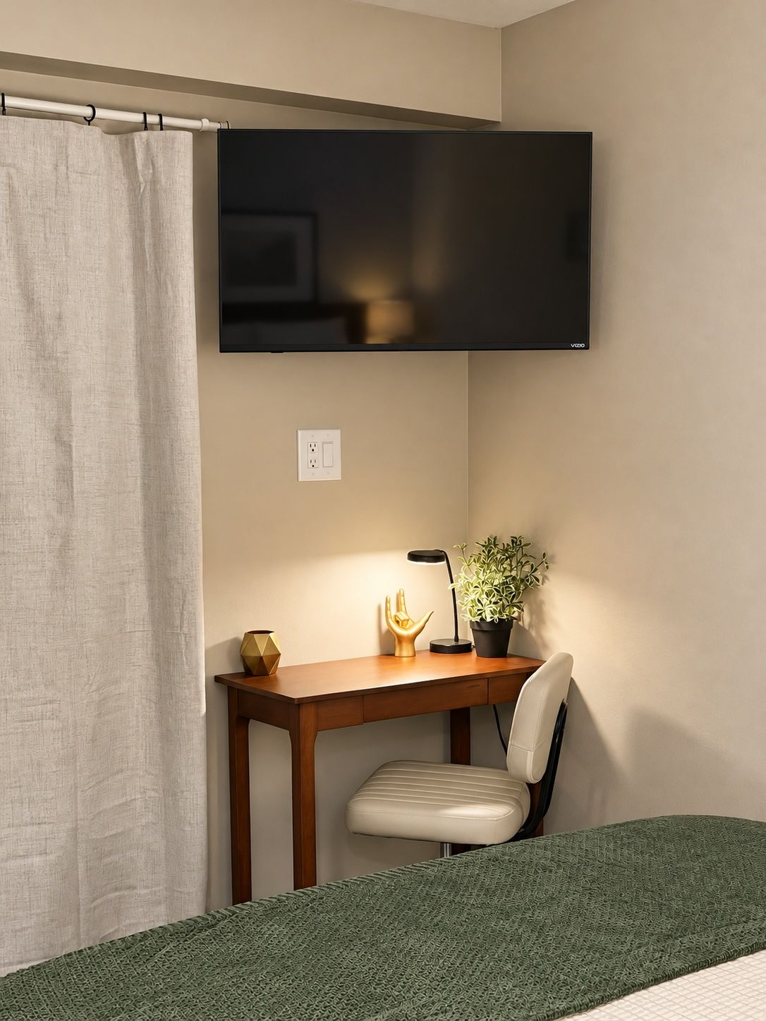 Une chambre dotée d’un téléviseur à écran plat fixé au mur, d’un bureau avec une chaise, d’une plante en pot et d’un lit recouvert d’une couverture verte.