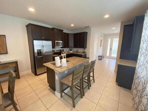 A modern kitchen with dark cabinetry, a central island with bar stools, and a stainless steel refrigerator. - Elegant 8BR Family Home w/ Pool & Spa (Kissimmee)
