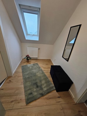 Una habitación pequeña y estrecha con techo inclinado, un radiador, una planta en maceta, una alfombra verde y un banco negro.