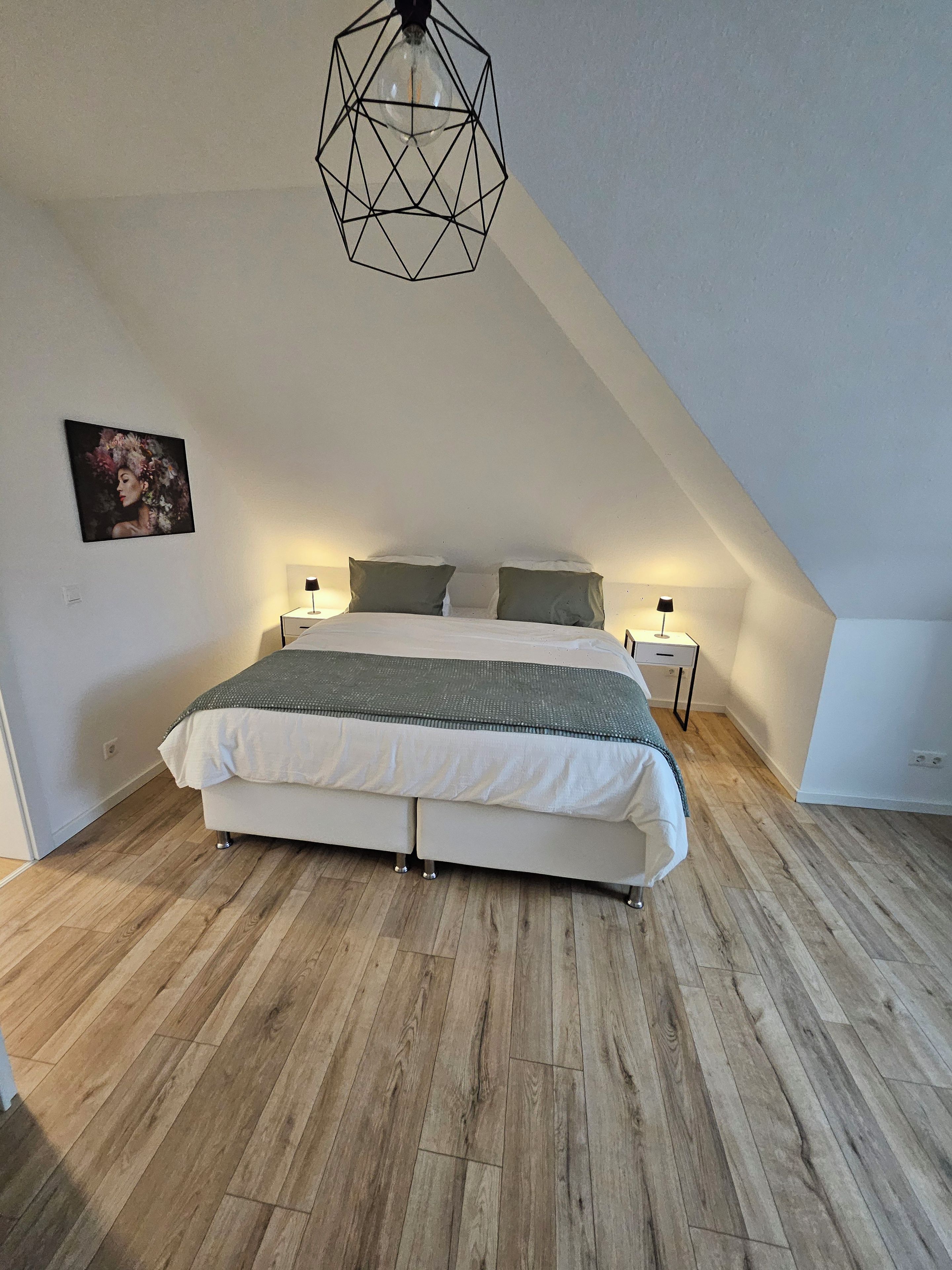Un dormitorio con techo inclinado, una cama con colcha blanca y gris, dos mesitas de noche con lámparas y una lámpara colgante de diseño geométrico.