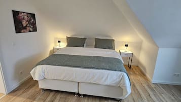 Un dormitorio con techo inclinado, una cama con colcha blanca y gris, dos mesitas de noche con lámparas y una lámpara colgante de diseño geométrico.