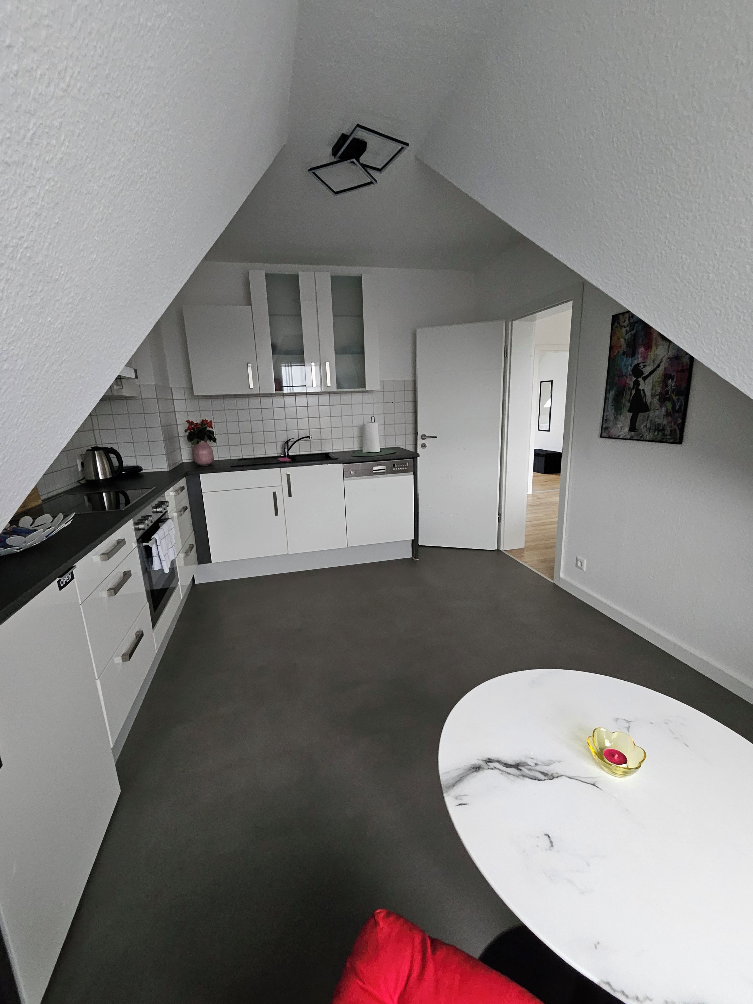 Une cuisine compacte avec des meubles blancs, un plan de travail sur lequel se trouve une bouilloire, et un coin repas avec une table et un coussin rouge.
