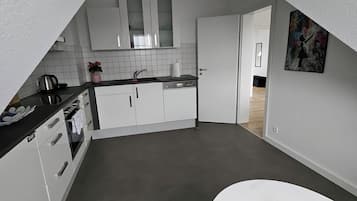 Une cuisine compacte avec des meubles blancs, un plan de travail sur lequel se trouve une bouilloire, et un coin repas avec une table et un coussin rouge.