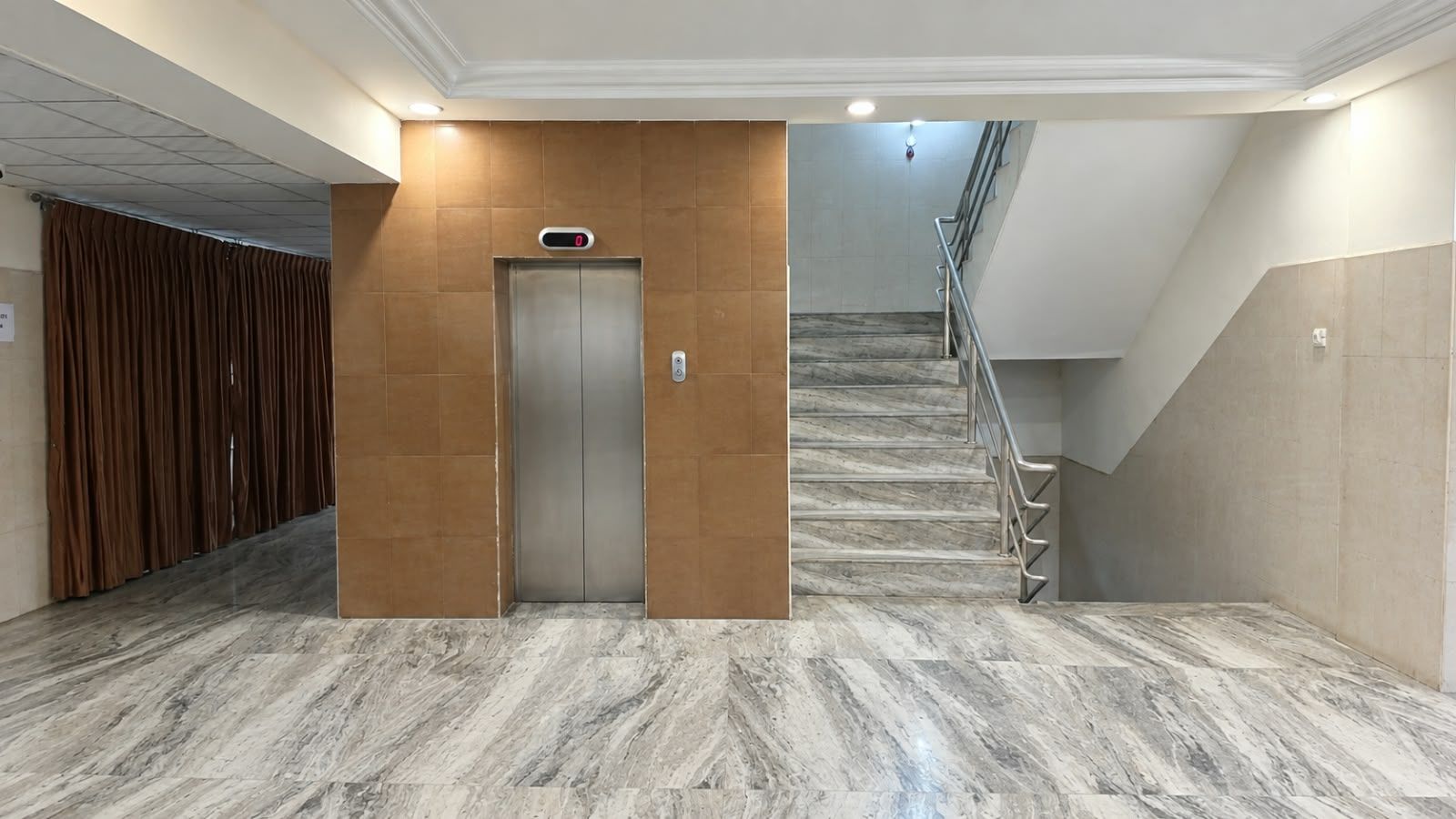 An elevator in a hallway with a marble floor, a staircase with metal railings, and a wooden panel on the wall.