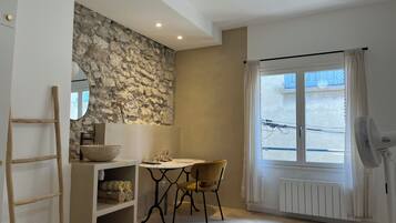 Een kamer met een stenen muur, een houten ladder, een wastafel, een tafel met een stoel, een raam met gordijnen en een staande ventilator.