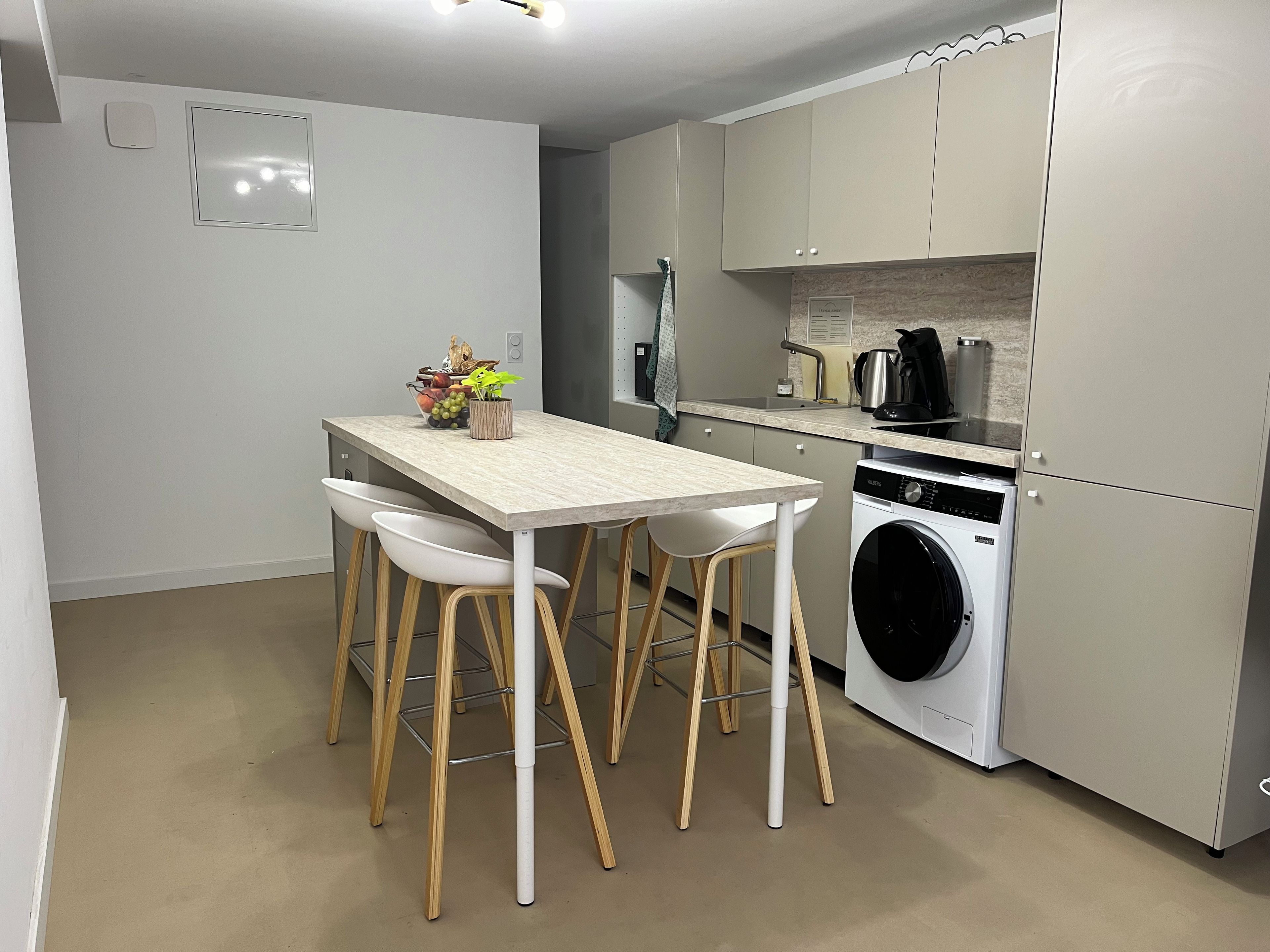 Een compacte keuken met een wasmachine, een koffiezetapparaat en een klein eethoekje met krukjes.