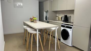 Een compacte keuken met een wasmachine, een koffiezetapparaat en een klein eethoekje met krukjes.