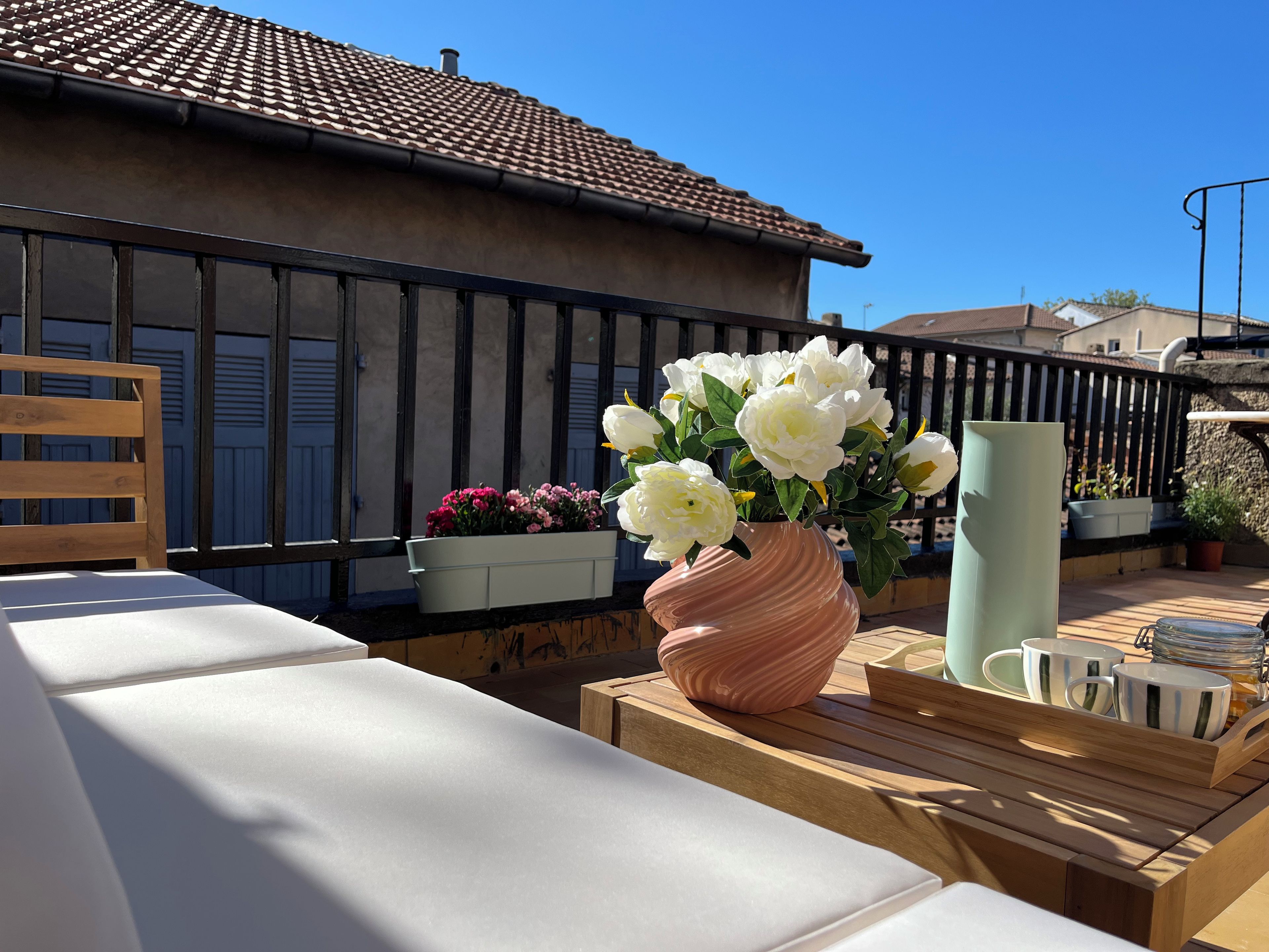 Een balkon met een houten bankje, een roze vaas met witte bloemen en een metalen hekwerk.