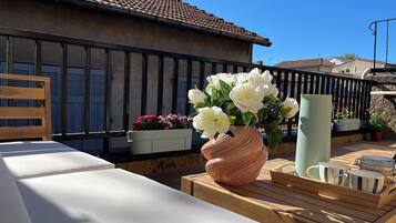 Een balkon met een houten bankje, een roze vaas met witte bloemen en een metalen hekwerk.