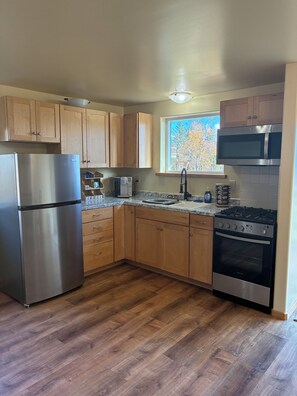 A kitchen with wooden cabinets, a stainless steel refrigerator, a microwave, a gas stove, and a window with a view of trees. - Experience Alaska - Relaxing cottage on 9 acres with glacier and ocean views (Fritz Creek)