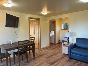 A room with a dining table, chairs, a blue sofa, a TV mounted on the wall, and a laundry area with a washing machine. - Experience Alaska - Relaxing cottage on 9 acres with glacier and ocean views (Fritz Creek)