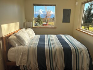A bedroom with a bed, a window with a view of trees and mountains, and a wall-mounted air conditioning unit. - Experience Alaska - Relaxing cottage on 9 acres with glacier and ocean views (Fritz Creek)