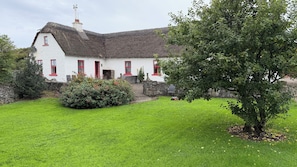 A white house with red trim and a thatched roof, surrounded by a well-maintained lawn and a large tree. - 3 bed 3 bath cottage; Minutes from Cliffs of Moher (CE)
