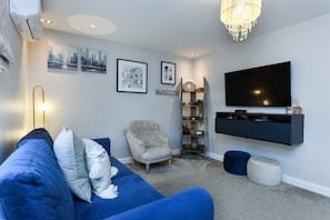 A modern living room with a blue sofa, a tufted armchair, a mounted TV, and a chandelier. - 2-Bed Home with Parking Near Trent Bridge (Nottingham)