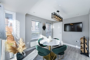 A modern dining area with a round table, green upholstered chairs, a pendant light, and a flat-screen TV mounted on the wall. - 2-Bed Home with Parking Near Trent Bridge (Nottingham)