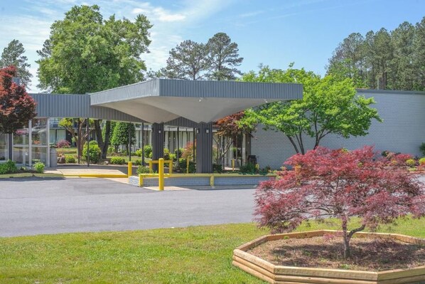 A modern building with a covered entrance, a landscaped garden with trees, and a paved driveway.