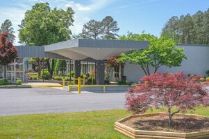 A modern building with a covered entrance, a landscaped garden with trees, and a paved driveway.