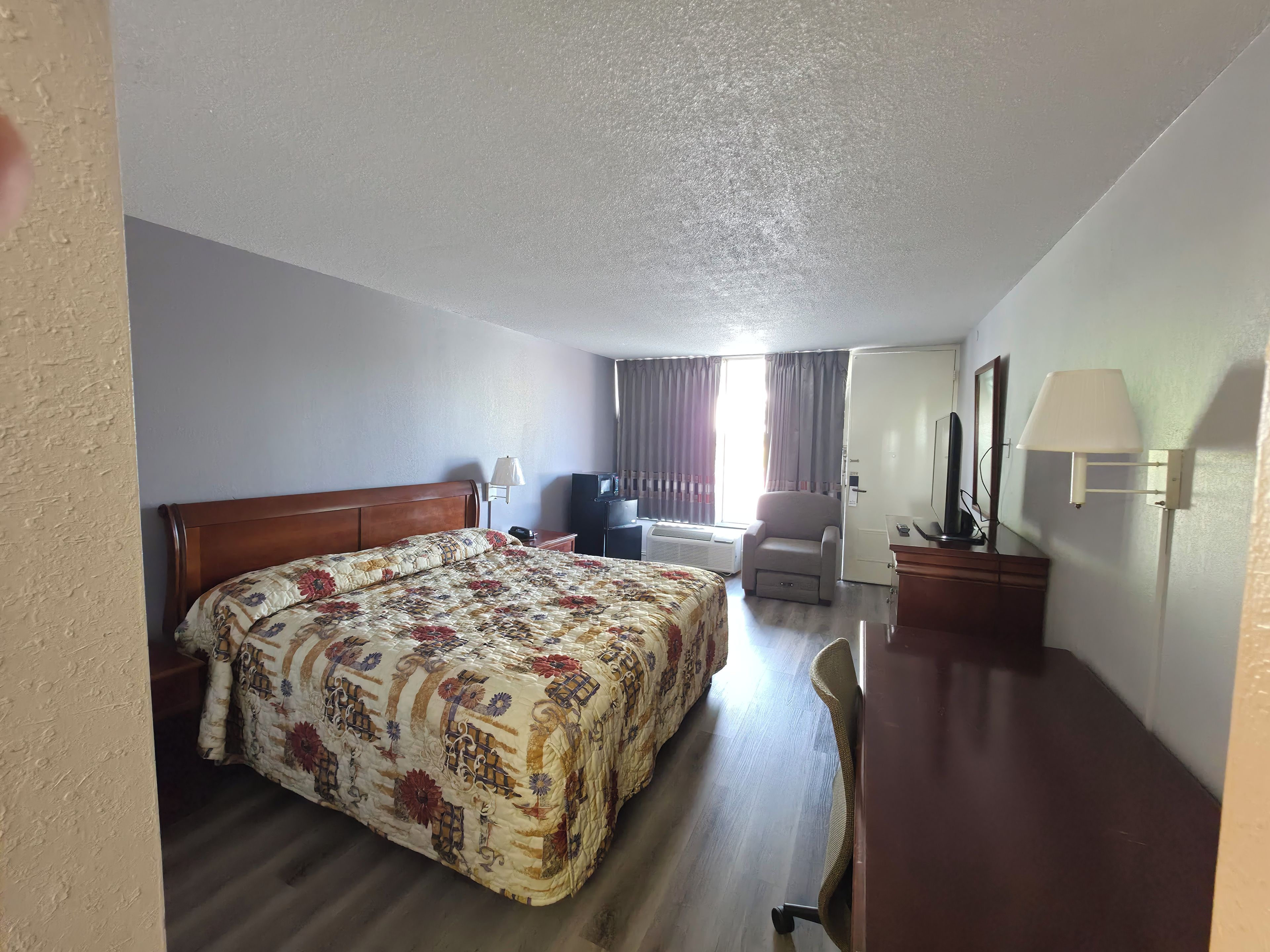Una habitación de hotel con cama, escritorio, silla, lámpara, televisor y ventana con cortinas.