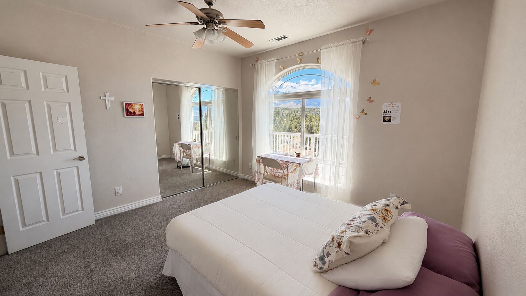 A bedroom with a bed, a ceiling fan, a window with curtains, and a balcony.