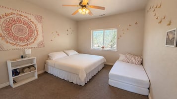A bedroom with a large bed, a smaller bed, a bookshelf, a window, and a ceiling fan.
